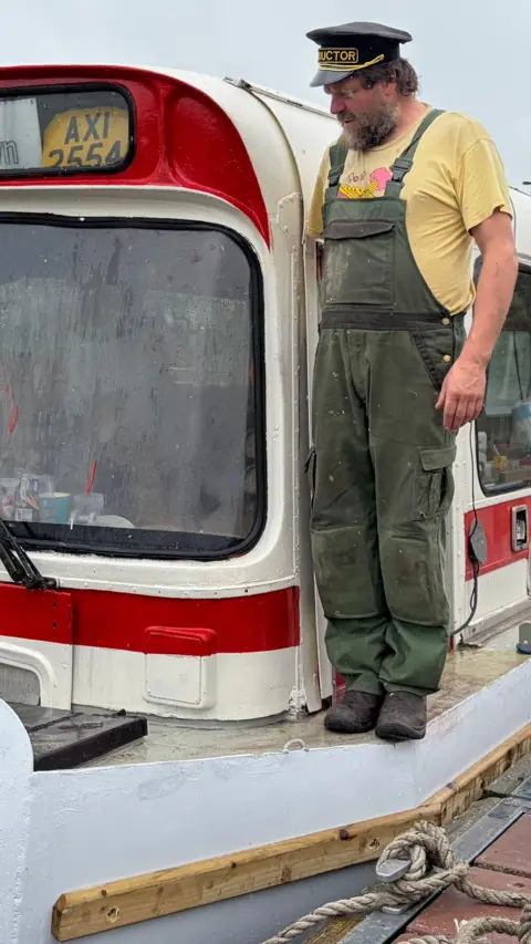 Man wearing dungarees is standing on a boat made from an old bus 