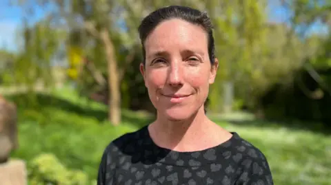 President of Policy and Resources, Deputy Lindsay De Sausmarez. She has short dark hair which is tied back. She is looking into the camera. She is wearing a black jumper with grey hearts on it. The background is a blurred image of a grassy area.