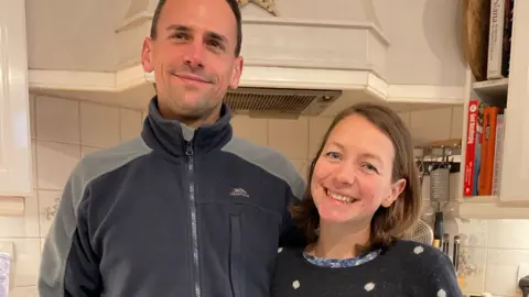 A man wearing a dark fleece and a woman with a spotted sweater in a kitchen.