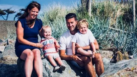 Jennie Bailey Two adults and two young boys sat on some rocks on a beach, with blue sky and a grassy verge behind them. The woman has long dark hair and wears a navy dress. The man has short dark hair and a white t-shirt. Both toddlers are blonde and are wearing t-shirts and shorts. All four are looking at the camera and smiling.
