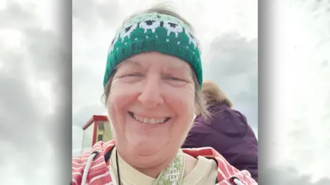 Supplied A woman with a green and white woolly headband, pink stripped hoodie, yellow t-shirt and a beige bag strap across her shoulder. Behind her is a woman in a purple coat and a cloudy sky.