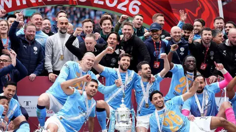 Manchester City players and Pep Guardiola celebrate with the FA Cup trophy following their side's victory over Chelsea at Wembley Stadium. The players are in pale blue and white and each has an arm in the air. Others - coaches, fans and others - are behind them, some also holding one arm in the air. Everyone is smiling.