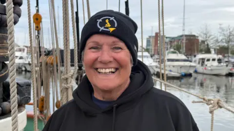 The photo shows a woman smiling at the camera. She's wearing a black beanie hat and a black hoodie. She's standing on a boat, and there are various ropes behind her. You can also see other boats in the marina in the background. 