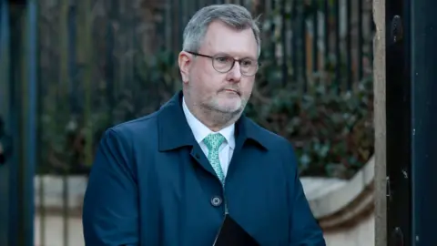 Jeffrey Donaldson, with short grey hair, wearing a white shirt, blue tie, navy jacket and glasses. He is holding a notebook. 