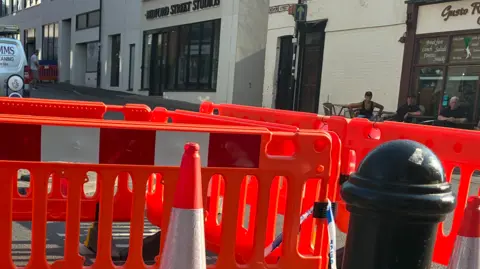 A cordoned-off area featuring barriers and a traffic cone is in the foreground. A building is in the background on Bedford Street.