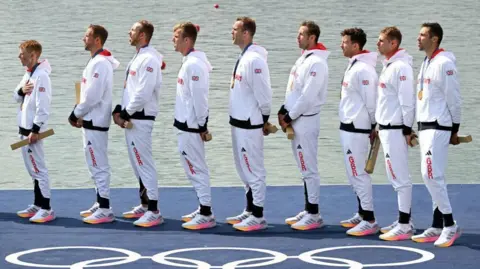 Great Britain's gold medallists listen to the national anthem on the podium during the medal ceremony after the men's eight final rowing competition at Vaires-sur-Marne Nautical Centre during the Paris 2024 Olympic Games. They stand in a row, facing left, in white tracksuits with the Union Jack emblazoned on their arms. They hold long, rectangular golden gift boxes and wear their gold medals.