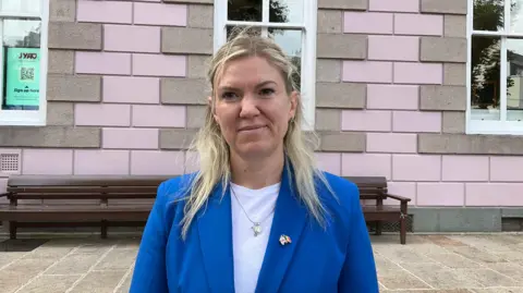 Deputy Raluca Kovacs. She is wearing a white top and a blue blazer. She is looking into the camera.