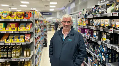 Mark Cox, a man with white hair, glasses and a grey beard. He is standing in a supermarket aisle, with pet food to the right of the image and cooking supplies - including olive oil and seasoning - to the left. He is wearing a 'coop' branded jacket with a zip through the middle, over a pink shirt.