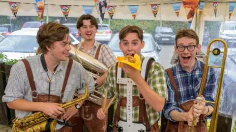 Anthony Upton / PA Media Four members of a band, holding musical instruments, with one band member seemingly about to eat a hot dog. They appear to be in a gazebo with clear sides, looking out onto a car park.