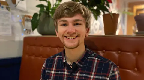 BBC Sam Braham has short brown hair and is wearing a checkered shirt. He is sitting on a brown sofa 