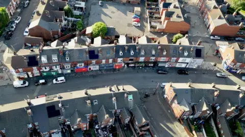 An aerial view of a residential neighbourhood with rows of closely packed houses and a small commercial strip along a street. The buildings are mostly made of red brick and feature grey or brown tiled roofs, many with chimneys evenly spaced along the rooftops. The houses appear to be terraced, sharing walls, and are arranged in parallel rows with narrow back gardens visible behind some of them.