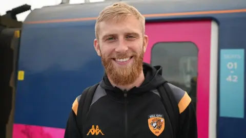 Jasmine Lowe / BBC Oliver McBurnie who has red hair and beard and is wearing a black Hull City top stands in front of the London train 