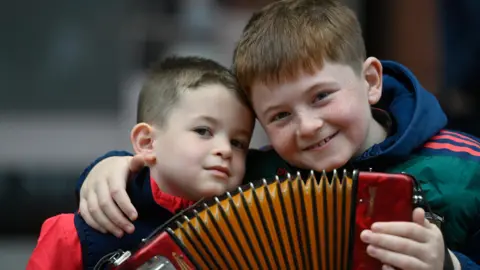 Two young boys with short brown hair sitting beside each other. They are holding an accordion. 
