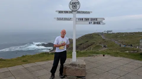 Handout Phil Brown at Land's End with a sign next to him and the headland and ocean behind him. He is wearing a white t-shirt with a motif on it and dark trousers.
