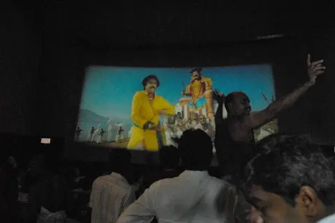The India Today Group via Getty Images Cheering fans watching a Rajinikanth film in a theatre in southern India. He can be seen on the screen wearing a yellow kurta. 