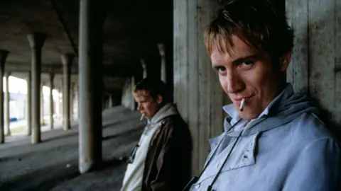 Moviestore Collection Ltd Two men with blond hair both smoking a cigarette standing underneath a motorway flyover with pillars in the background holding up the road deck