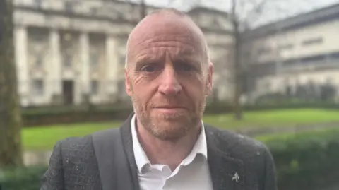 A close-up shot of James Brannigan. He is an older man with short, buzzed hair on his head. He has a light coloured beard. He is looking into the camera with a neutral expression on his face. He is standing outside a a court and is wearing a grey suit jacket and a white shirt.