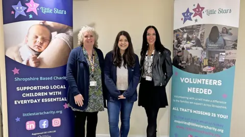 Housing Plus Group Three women standing together between two tall banners. The woman on the left has white blonde curly hair, a black, white and green dress and a navy blue cardigan. In the middle is Leanne, who has long brown hair, a white top and navy zip hoodie. On the right the final woman has long black hair, a zebra print top and a black leather jacket on. The banner on the left is purple and says "little stars, Shropshire based charity, supporting local children with everyday essentials". On the right, the banner is white and light blue, and reads "volunteers needed, all that's missing is you"