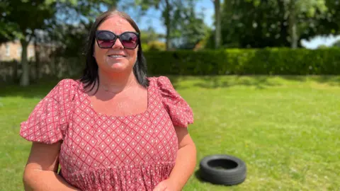 Jules stands outdoors on a sunny day, wearing a red patterned dress with short sleeves and holding an object in their hands. In the background, there is green grass, trees, and a black tire lying on the ground.