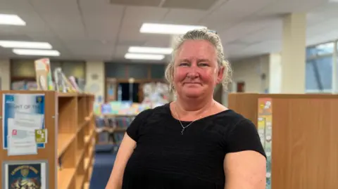 Claire Judd is standing indoors and facing the camera with a smile. She is wearing a black top and her fair hair is tied back. There are rows of library books behind her.