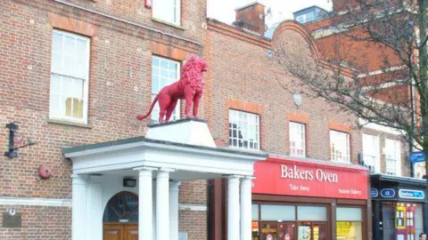 Colin Smith/Geograph A Red Lion statue on top of a canopy, on a white plinth, with shops to the right. The building is made from red brick with large windows. 