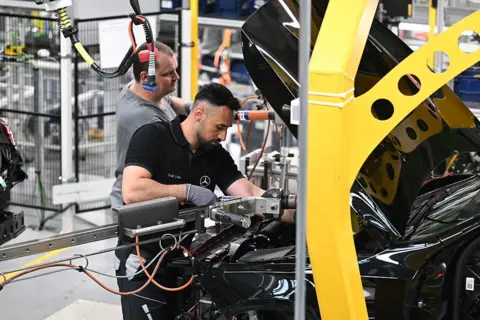 Getty Images Factory factor peers under the black vehicle cover and holds a mechanical arm in place while he is working