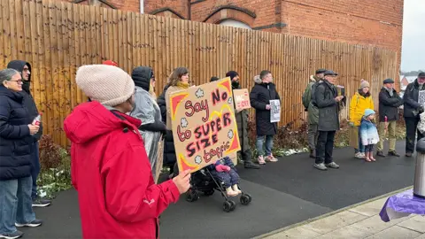 A group of people gather to protest against plans for the new McDonalds. One woman in a red coat holds a sign which reads "Say No To Supersize Traffic".