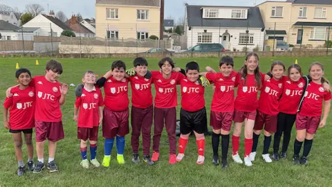 Jersey Bulls Foundation A row of children smiling with arms around each other all wearing red JTC logo'd football shirts, standing on a field. Behind is a road with parked cards and a few houses in a row. The skies are grey.
