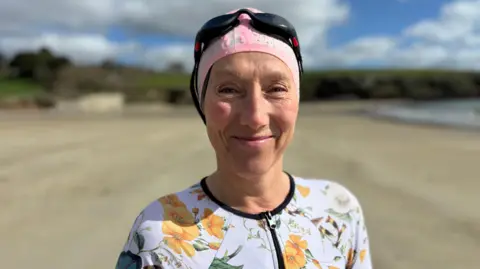 A woman wearing a white flowery swimming costume, a swimming hat and goggles