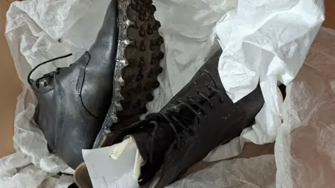 Mountain Heritage Trust Mabel Barker's hobnail boots from 1925. The pair of black leather lace-up boots have metal soles with small prongs fixed to them. They are wrapped up in white tissue. 