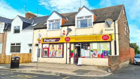 Darren Stanford A two-storey corner terrace brick house has been converted into a convenience store. There is a bus stop and a bin outside. The yellow sign above the shop reads: "Pallion Convenience Store" and has red Post Office logos on it too. The shop windows are dotted with colourful posters, and there is an ATM on the left.