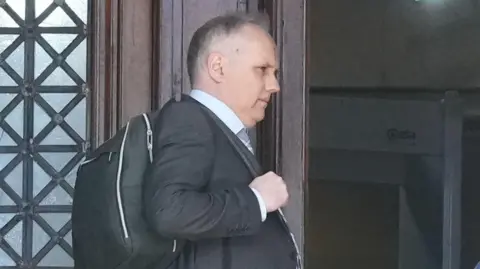 Dylan Dawes standing leaning on a door. He has a bag over his shoulder. 