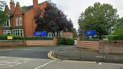 The entrance of the Priory Academy LSST in Lincoln with two blue signs either side of the gate which is behind a low red-brick wall with a house on one side and large trees and a hedge running behind the brick wall