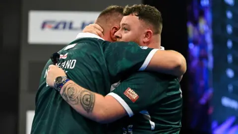 Two men wearing a green sports top hugging as they won the championship. Both have short dark hair, one is holding darts in his hand, has a silver watch on his wrist and has tattoos up his arm.