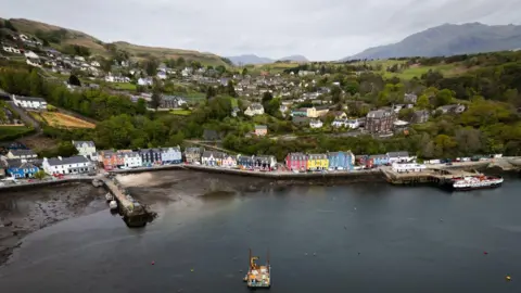 An aerial view of the Isle of Mull, including the port, with brightly coloured buildings at the sea front and sprawling hills in the distance