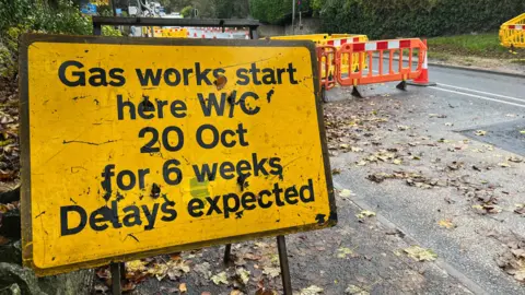 Close up of yellow roadworks information sign, reading: "Gas works starts here W/C 20 Oct for 6 weeks. Delays expected". In the background, there are orange barriers and cones on a wide road. 
