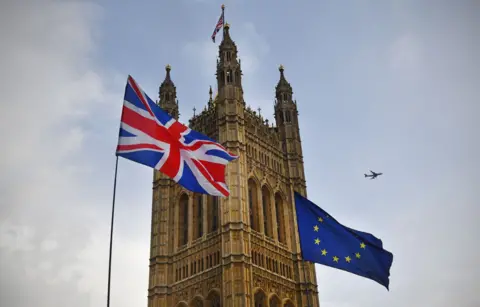 BBC News A union flag and the flag of Europe are blowing in the breeze in front of the Houses of Parliament. The sky is blue with clouds and an aeroplane can be seen in the distance. 