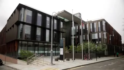 The council headquarters. A modern three-storey building with ceiling-to-floor windows. There are three flag poles on the pavement outside.