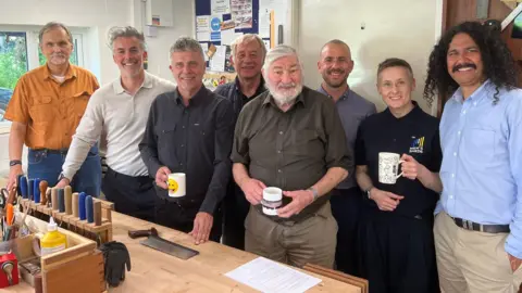 A group of eight people - seven men and one women - standing in a shed and smiling at the camera. Three of them are holding mugs of tea. They are standing behind a workbench, which has tools set out tidily on it.