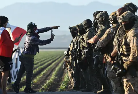 Getty Images Two protesters in dust masks use their phones to film a line of heavily armed National Guard troops standing in a field of crops in California farmland