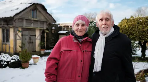 ITV News West Country The Buntings standing in the snow in their garden, dressed up warmly, with one of the replica houses, the timber-framed Elizabethan house, in the background