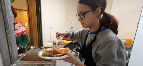 A girl wearing glasses and a grey sweatshirt pours baked beans over two slices of white toast.