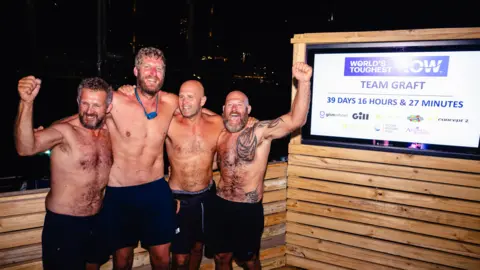 World's Toughest Row Four men are standing bare chested and wearing black shorts in front of a sign which gives their time as 39 days, 16 hours and 27 minutes.