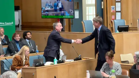 New leader Ian Thorn (left) shakes hands with former leader, Conservative Richard Clewer (right) while in the council chamber. They have other councillors sitting around them in a space that is a little like a lecture theatre with lots of wooden panelling 