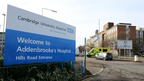 A large blue and white sign at the entrance to Addenbrooke's Hospital, advising people they have arrived. A car and an ambulance are being driven along a road in the background.