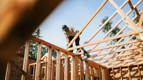 A builder fitting the wooden frame of a house together. The man is standing on top of the frame and is slotting a wooden beam into place. He is wearing a t-shirt and cap.