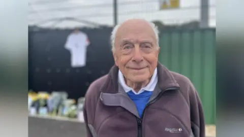 George Carden/BBC Michael Stemp posó para una foto en el fútbol Chichester FC. Michael llevaba una camisa blanca, un jersey azul y una chaqueta con cremallera negra. Tiene el pelo blanco. El tributo floral y de la tarjeta se puede ver en el fondo, aunque parcialmente borrosa.