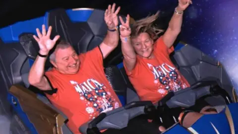 Supplied Phil Taylor, who has short grey hair and is wearing a red t-shirt, and Melanie Taylor, who has long blonde hair and red t-shirt, have their arms up in the air while sitting in a blue and grey roller coaster. It's clearly moving, as Melanie's hair is being swung around.