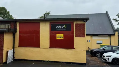 A single storey building with bricks painted in pale yellow with boarded up windows painted burgundy. A sign says 'Educ8 Alternative Education School' and a sign below says 'warning' and 'hardhat'.