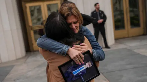 Reuters A woman with blonde hair looks distraught as she hugs another mum, who has black hair, while holding a framed photograph of her son in his school uniform outside a court in Los Angeles.